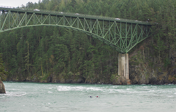 Kayakers paddling through large waves underneath Deception Pass Bridge